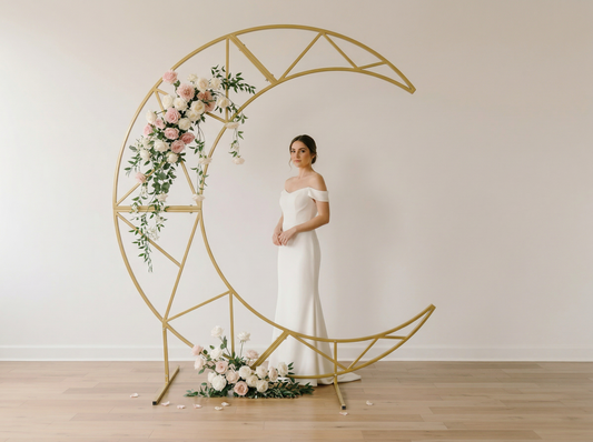 A bride standing next to a decorative gold metal crescent wedding arch with floral arrangements