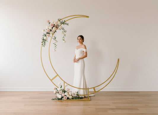 Woman in a white wedding dress standing next to a decorative gold arch with flowers against a plain wall.