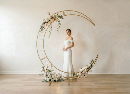 Woman in a white dress standing inside a decorative floral arch against a plain wall.